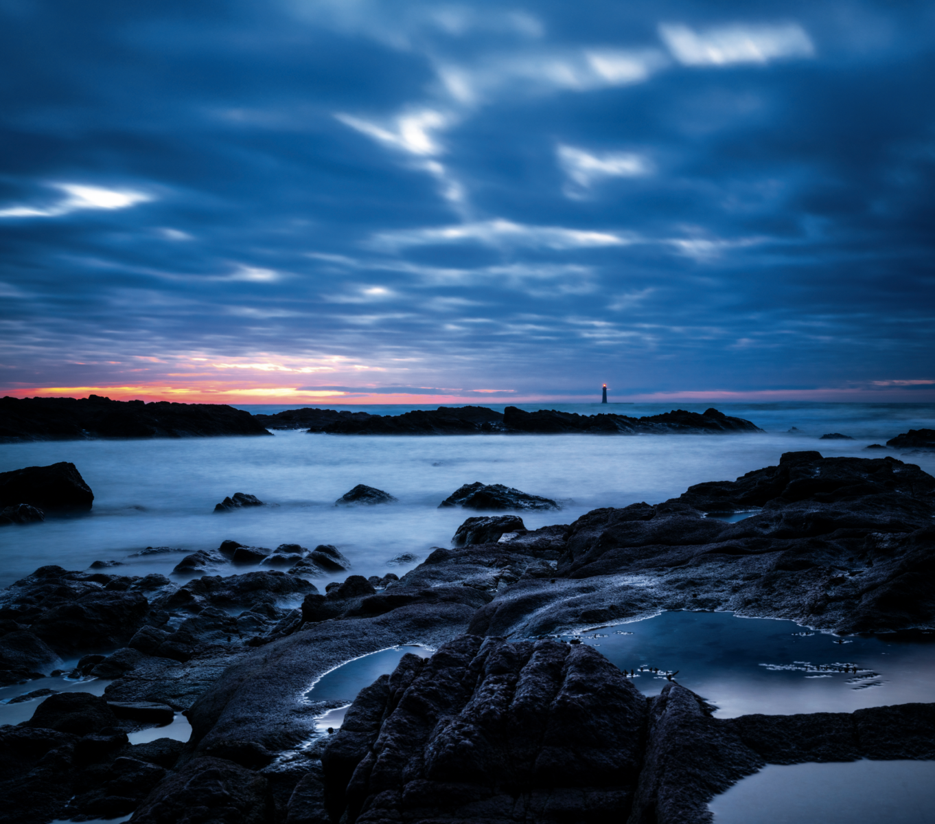 Vue de la côte rocheuse des Sables d'Olonne lors d'un couché de soleil rose et orange qui perse à peine dans un ciel très nuageux. Au loin, le phare des barges brille dans la pénombre.