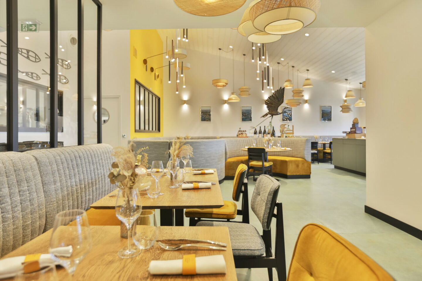 Intérieur d'un restaurant moderne avec des tables en bois, des sièges rembourrés gris, des accents jaunes, des lampes suspendues et un décor minimaliste. Les tables sont garnies de verres et de serviettes, ce qui crée une atmosphère chaleureuse et accueillante. - Édition Offset 5 (Partenaire de votre communication locale en Vendée)