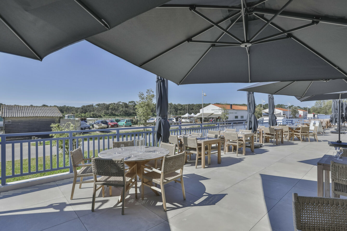 Terrasse du restaurant en plein air avec tables en bois et chaises en osier sous de grands parasols noirs, donnant sur un port de plaisance avec des bateaux et des bâtiments au bord de l'eau par une journée ensoleillée. - Édition Offset 5 (Partenaire de votre communication locale en Vendée)