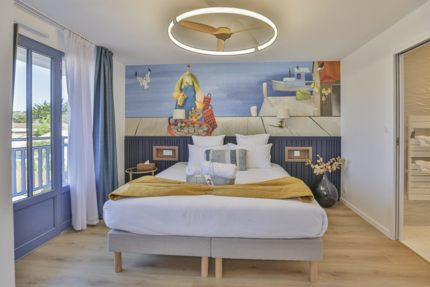 Une chambre à coucher moderne avec un grand lit, une couverture jaune, des oreillers décoratifs, un tableau mural au-dessus de la tête de lit, du parquet, un plafonnier rond et une porte ouverte sur une salle de bains. Une fenêtre avec des rideaux bleus se trouve sur la gauche. - Édition Offset 5 (Partenaire de votre communication locale en Vendée)