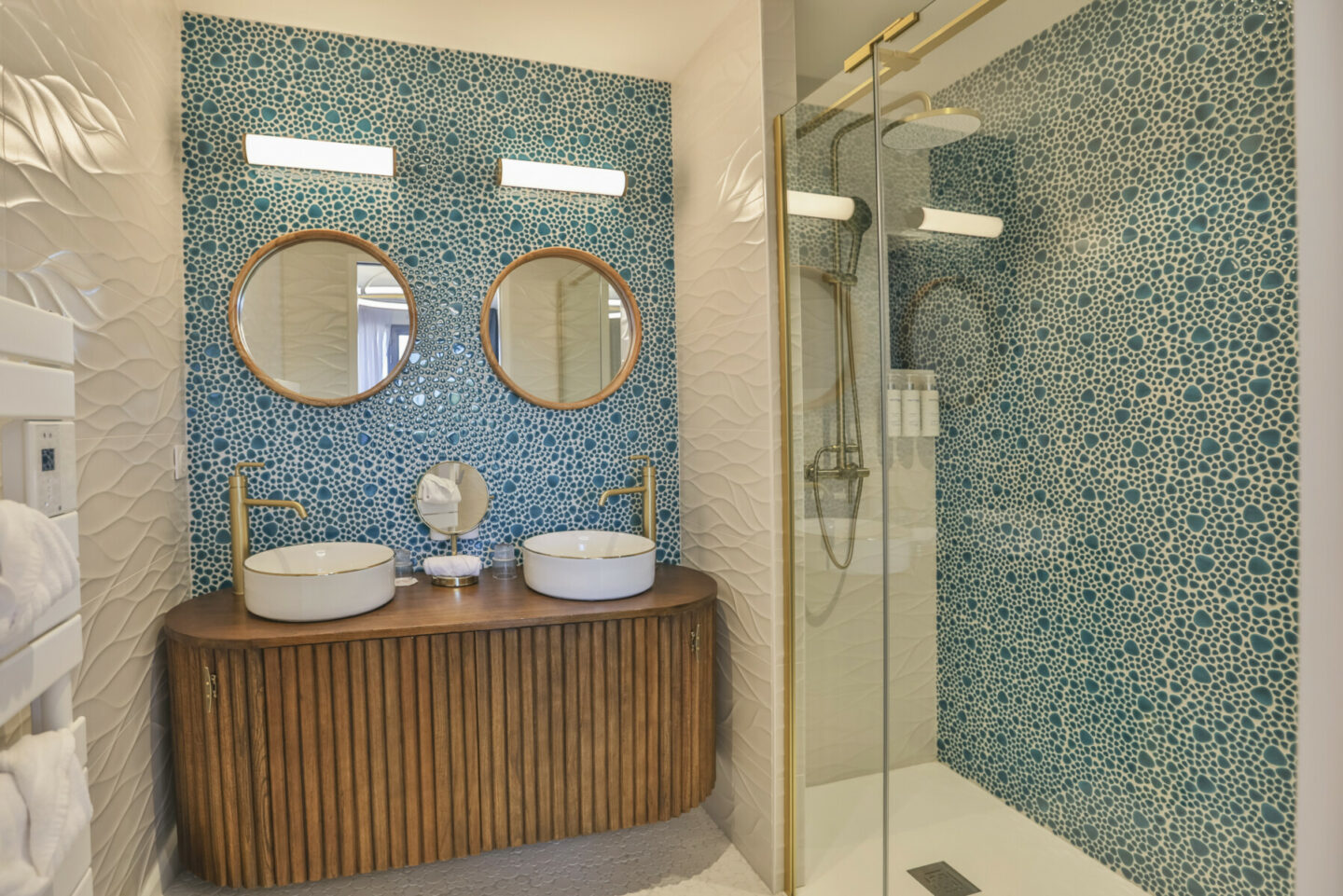 Salle de bains moderne avec un mur en mosaïque bleue, deux miroirs ronds au-dessus d'un meuble-lavabo en bois avec deux vasques et des robinets en or, et une douche en verre avec des carreaux en mosaïque bleue assortis et un appareil de douche en or. - Édition Offset 5 (Partenaire de votre communication locale en Vendée)