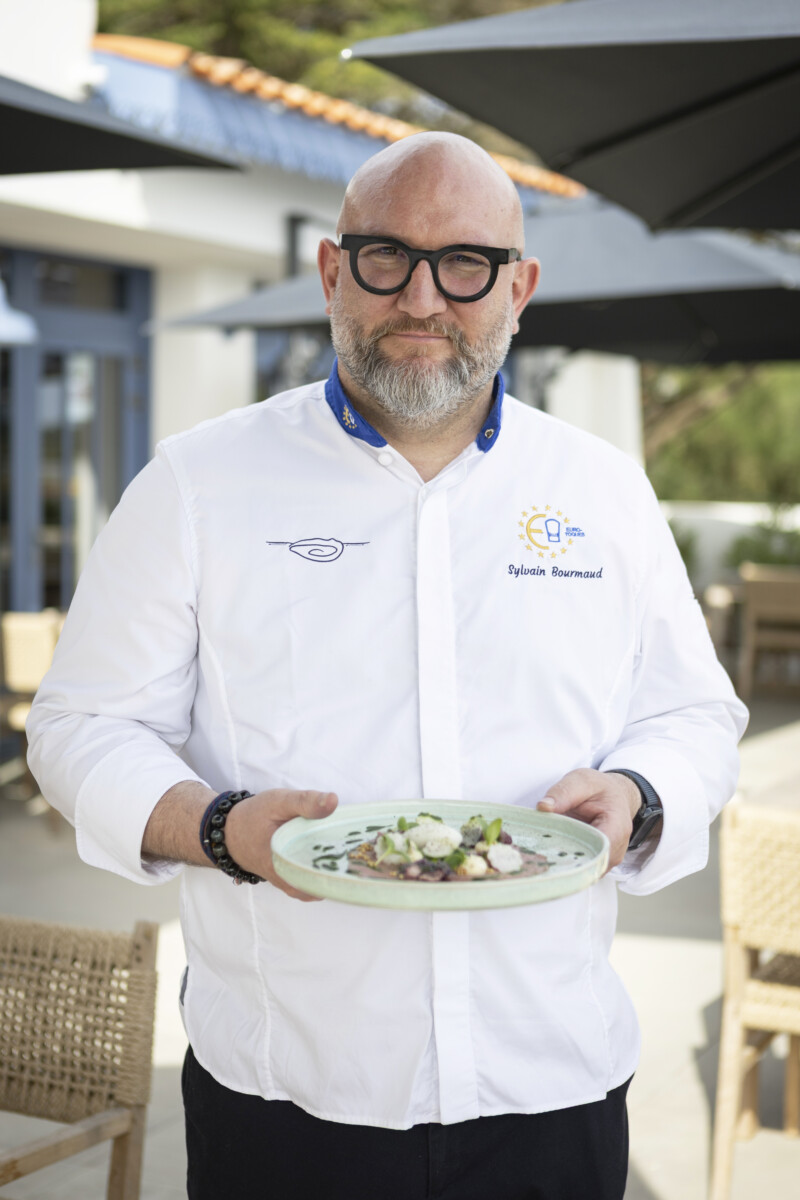 Un chef portant des lunettes et un uniforme blanc tient un plat en plein air dans le patio d'un restaurant. Il se tient devant des tables et des chaises, avec des parasols et un bâtiment visible à l'arrière-plan. - Édition Offset 5 (Partenaire de votre communication locale en Vendée)