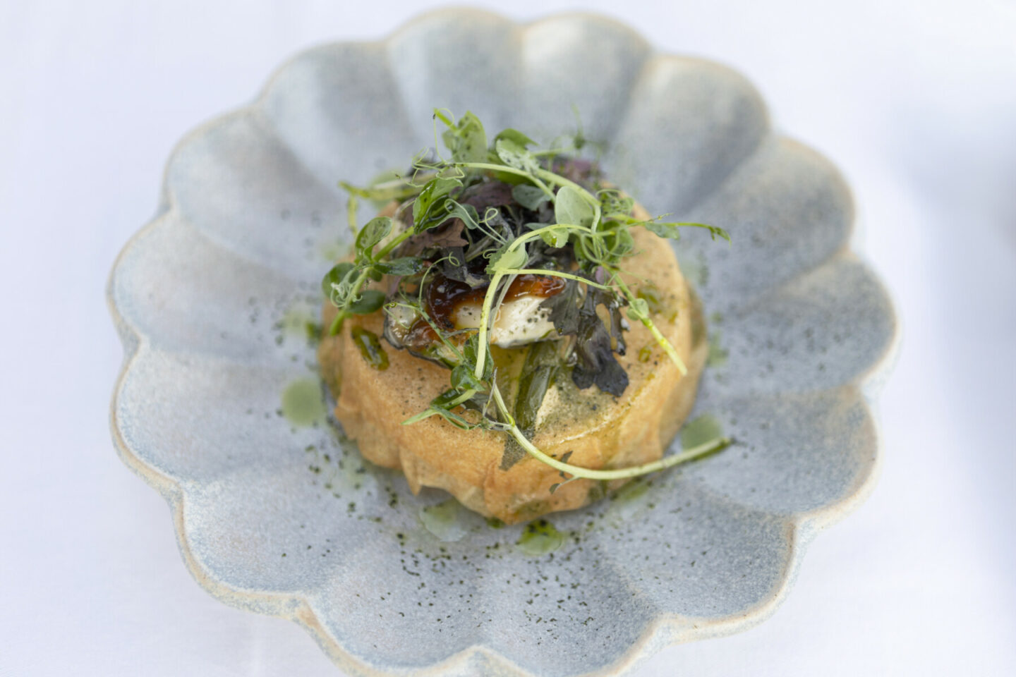 Un plat gastronomique composé d'une pâte salée garnie de fromage grillé et de micro-légumes frais, servi dans une assiette en céramique grise festonnée sur fond blanc. - Édition Offset 5 (Partenaire de votre communication locale en Vendée)