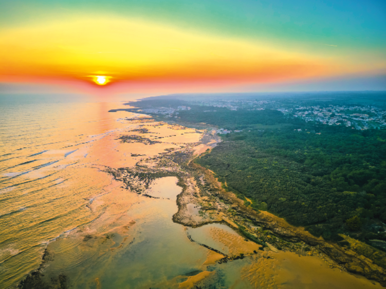 Vue aérienne d'un littoral au coucher du soleil, avec la lumière dorée du soleil se reflétant sur la mer et le rivage, une forêt verdoyante sur la droite, et une petite ville au loin - parfait pour vivre Les nocturnes du Château de Talmont sous un ciel coloré.