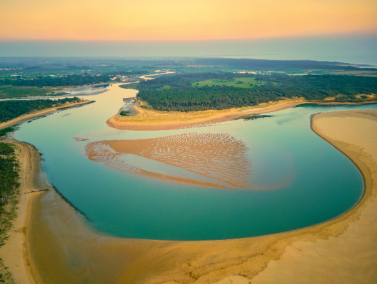 Vue aérienne d'une rivière sinueuse rencontrant un rivage sablonneux au coucher du soleil, avec des champs verts et des forêts en arrière-plan sous un ciel orange doux, rappelant la magie des nocturnes du Château de Talmont. - Édition Offset 5 (Partenaire de votre communication locale en Vendée)