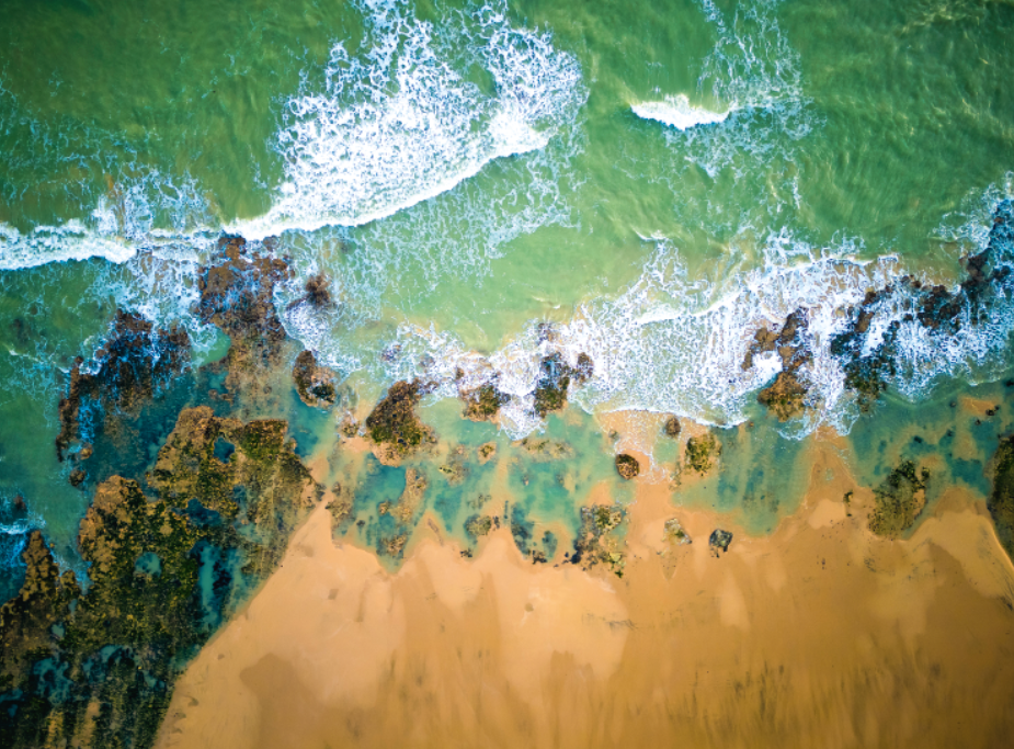 Vue aérienne des vagues s'écrasant sur une plage de sable avec des affleurements rocheux et une eau turquoise peu profonde, rappelant la beauté du littoral entourant Les nocturnes du Château de Talmont. - Édition Offset 5 (Partenaire de votre communication locale en Vendée)