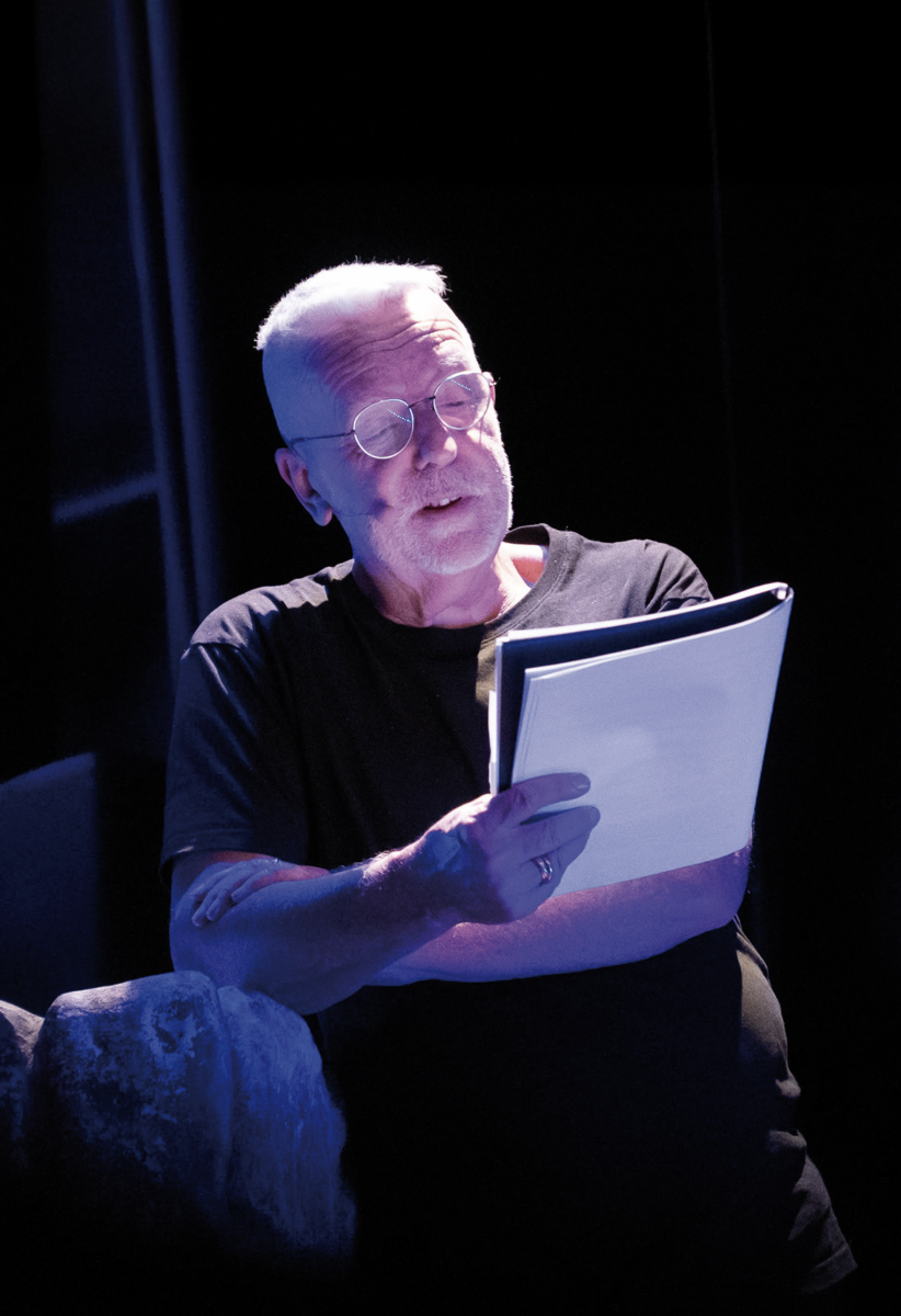 Un homme âgé aux cheveux blancs courts et aux lunettes rondes lit un texte sous un éclairage de scène dramatique, vêtu d'un t-shirt sombre et debout sur un fond sombre. - Édition Offset 5 (Partenaire de votre communication locale en Vendée)