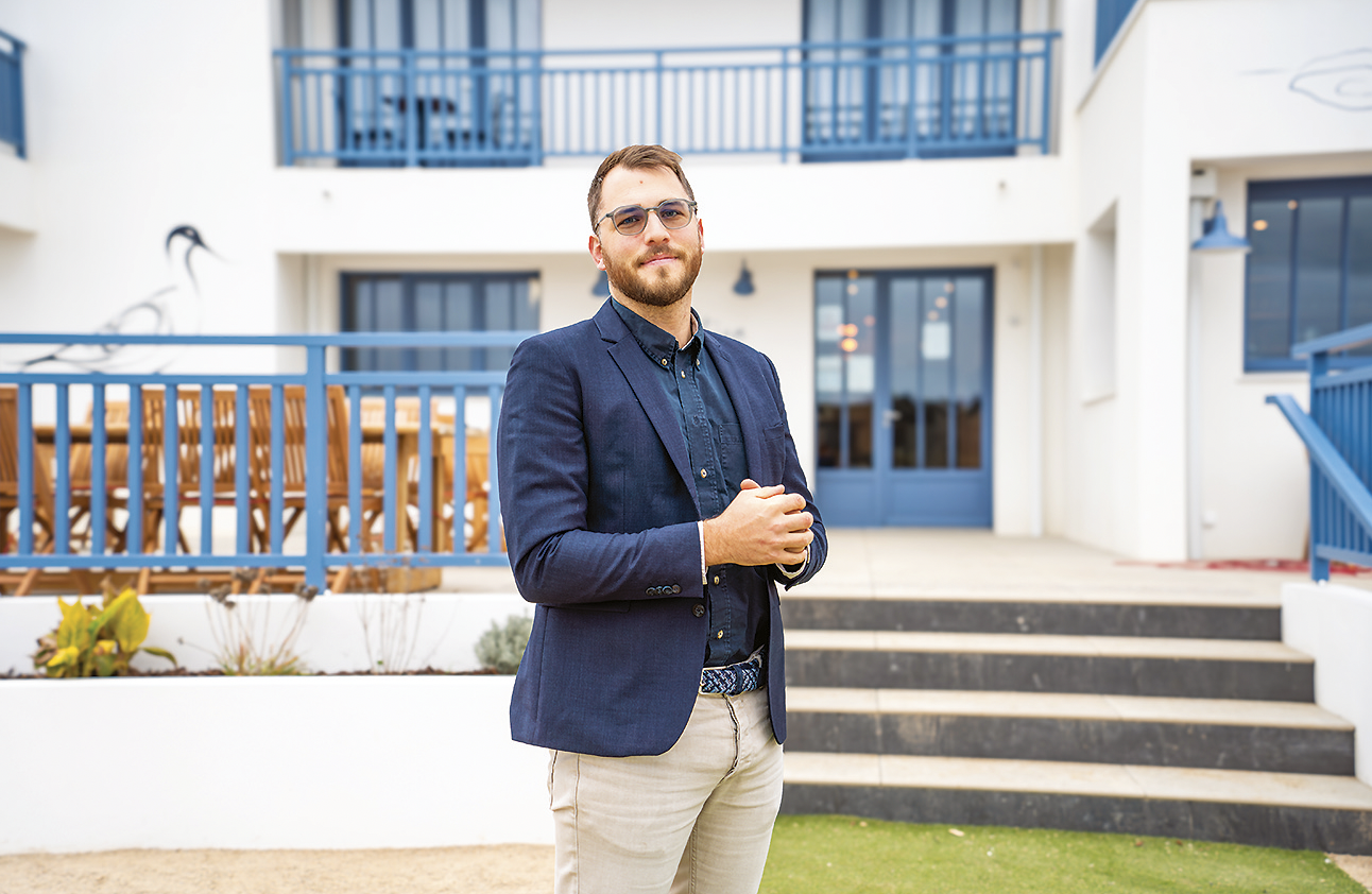 Un homme vêtu d'un blazer bleu marine, d'une chemise foncée et d'un pantalon clair se tient avec assurance à l'extérieur d'un bâtiment moderne blanc avec des accents bleus et des balcons, regardant la caméra les mains croisées. - Édition Offset 5 (Partenaire de votre communication locale en Vendée)