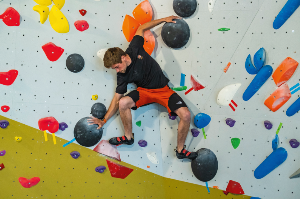 Un homme vêtu d'une chemise noire et d'un short orange escalade un mur de bloc intérieur à Veillon, en saisissant de grandes prises noires parmi des prises d'escalade colorées. - Édition Offset 5 (Partenaire de votre communication locale en Vendée)