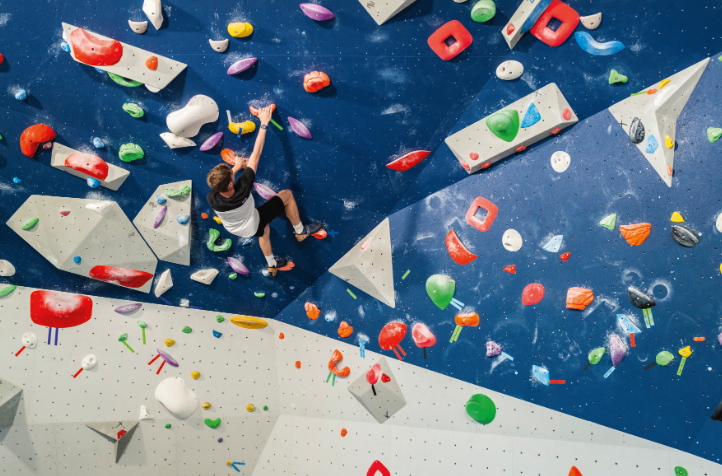 Une personne escalade un mur de bloc intérieur à Veillon, en s'agrippant à des prises colorées attachées à des surfaces bleues et blanches inclinées, vues d'en haut. - Édition Offset 5 (Partenaire de votre communication locale en Vendée)