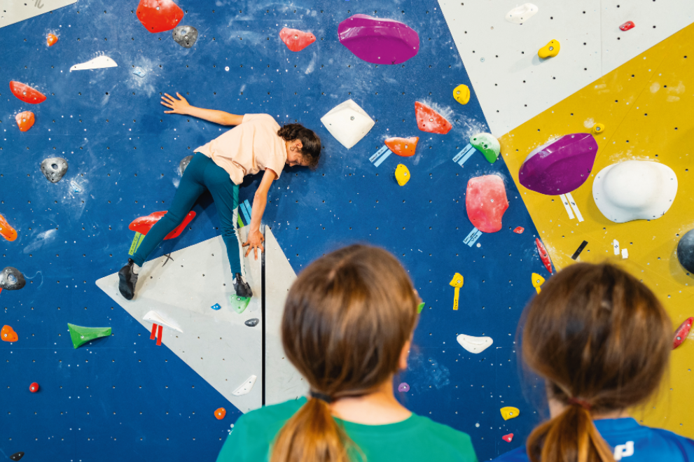 Un enfant escalade un mur d'escalade intérieur coloré, conçu avec des prises inspirées de Veillon dans des formes vives, tandis que deux autres enfants regardent d'en bas, dos à la caméra. - Édition Offset 5 (Partenaire de votre communication locale en Vendée)