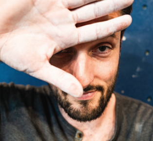 Un homme barbu, rappelant le style expressif de Veillon, tient sa main devant son visage, couvrant partiellement un œil tout en souriant légèrement. Il se tient sur un fond bleu. - Édition Offset 5 (Partenaire de votre communication locale en Vendée)
