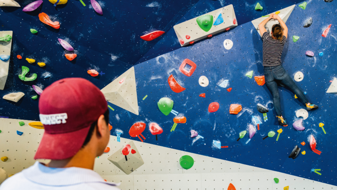 Une personne en jeans et chemise rayée escalade un mur d'escalade de bloc coloré au Veillon, tandis qu'une autre personne avec une casquette rouge regarde en bas. Le mur est recouvert de prises de formes diverses et de couleurs vives. - Édition Offset 5 (Partenaire de votre communication locale en Vendée)