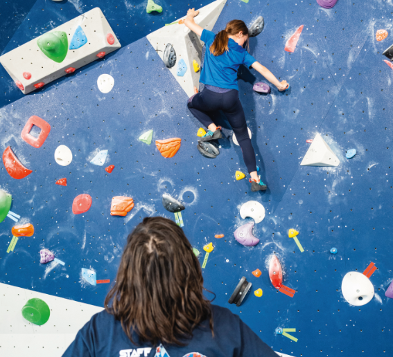 Deux personnes dans une salle d'escalade ; l'une escalade un mur Veillon bleu avec des prises colorées tandis que l'autre, vue de dos, regarde. Les deux personnes portent des chemises bleues et des vêtements de sport. - Édition Offset 5 (Partenaire de votre communication locale en Vendée)