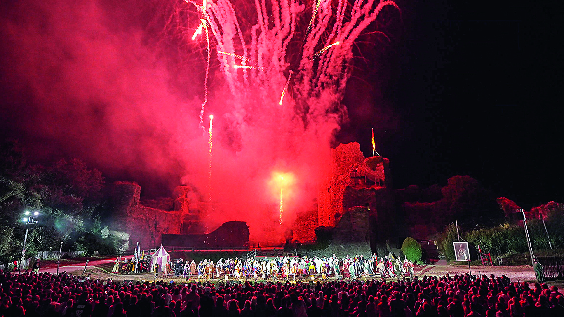 Une foule nombreuse assiste à une représentation sur scène devant des ruines anciennes, illuminées par des feux d'artifice rouge vif qui explosent dans le ciel nocturne.