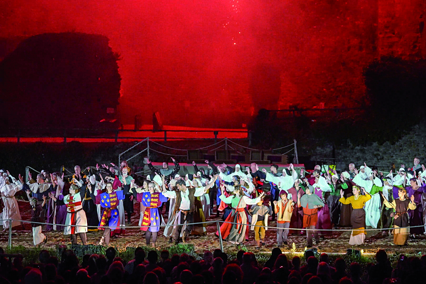 Des artistes en costumes médiévaux colorés reconstituent une scène historique en plein air, la nuit, sous le regard de la foule et l'éclairage rouge de la scène. - Édition Offset 5 (Partenaire de votre communication locale en Vendée)