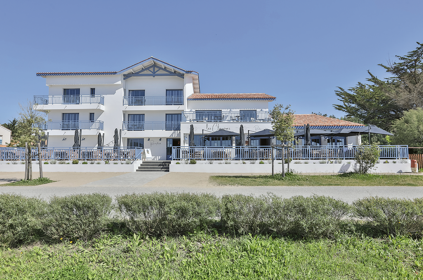 Un bâtiment moderne, blanc, de trois étages, avec des balustrades et des balcons bleus, se trouve sous un ciel bleu clair, entouré de verdure et d'arbres - idéal pour se détendre après avoir profité des nocturnes du Château de Talmont. Des marches mènent à un patio à l'entrée. - Édition Offset 5 (Partenaire de votre communication locale en Vendée)