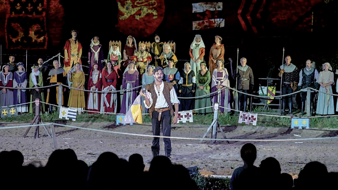 Un homme en costume médiéval se tient dans une arène, la nuit, et s'adresse à la foule. Des nobles et des royaux observent la scène depuis des sièges en gradins, sous des bannières héraldiques, évoquant des scènes présentées dans le magazine Talmont Saint-Hilaire. - Édition Offset 5 (Partenaire de votre communication locale en Vendée)