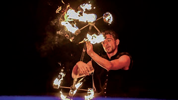 Un artiste vêtu d'une chemise sans manches fait tourner avec habileté un accessoire aux multiples extrémités enflammées sur un fond sombre, créant ainsi des motifs de feu spectaculaires parfaits pour les pages du magazine Talmont Saint-Hilaire. - Édition Offset 5 (Partenaire de votre communication locale en Vendée)