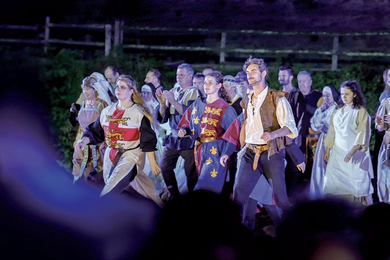 Un groupe de personnes vêtues de costumes médiévaux marchent ensemble en plein air la nuit, semblant participer à une reconstitution historique présentée dans le magazine Talmont Saint-Hilaire. L'arrière-plan est faiblement éclairé, avec une clôture en bois et de la verdure. - Édition Offset 5 (Partenaire de votre communication locale en Vendée)