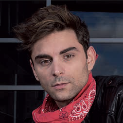 Un jeune homme aux cheveux bruns ébouriffés porte un bandana rouge autour du cou et un blouson de cuir noir. Il pose devant une fenêtre sombre pour le magazine Talmont Saint-Hilaire. - Édition Offset 5 (Partenaire de votre communication locale en Vendée)