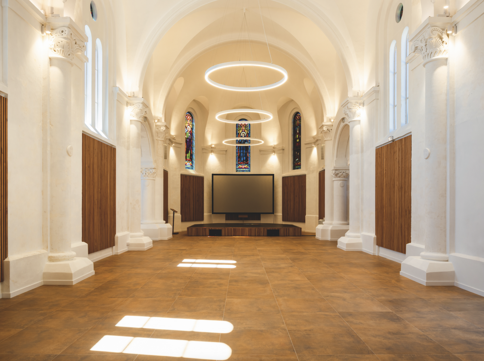 Une salle spacieuse et modernisée accueille les événements de Grimpe Atlantique Ouest, avec des plafonds voûtés, de grandes lampes circulaires suspendues, des vitraux et une scène avec un grand écran. Le sol carrelé et les panneaux muraux en bois créent une atmosphère accueillante. - Édition Offset 5 (Partenaire de votre communication locale en Vendée)