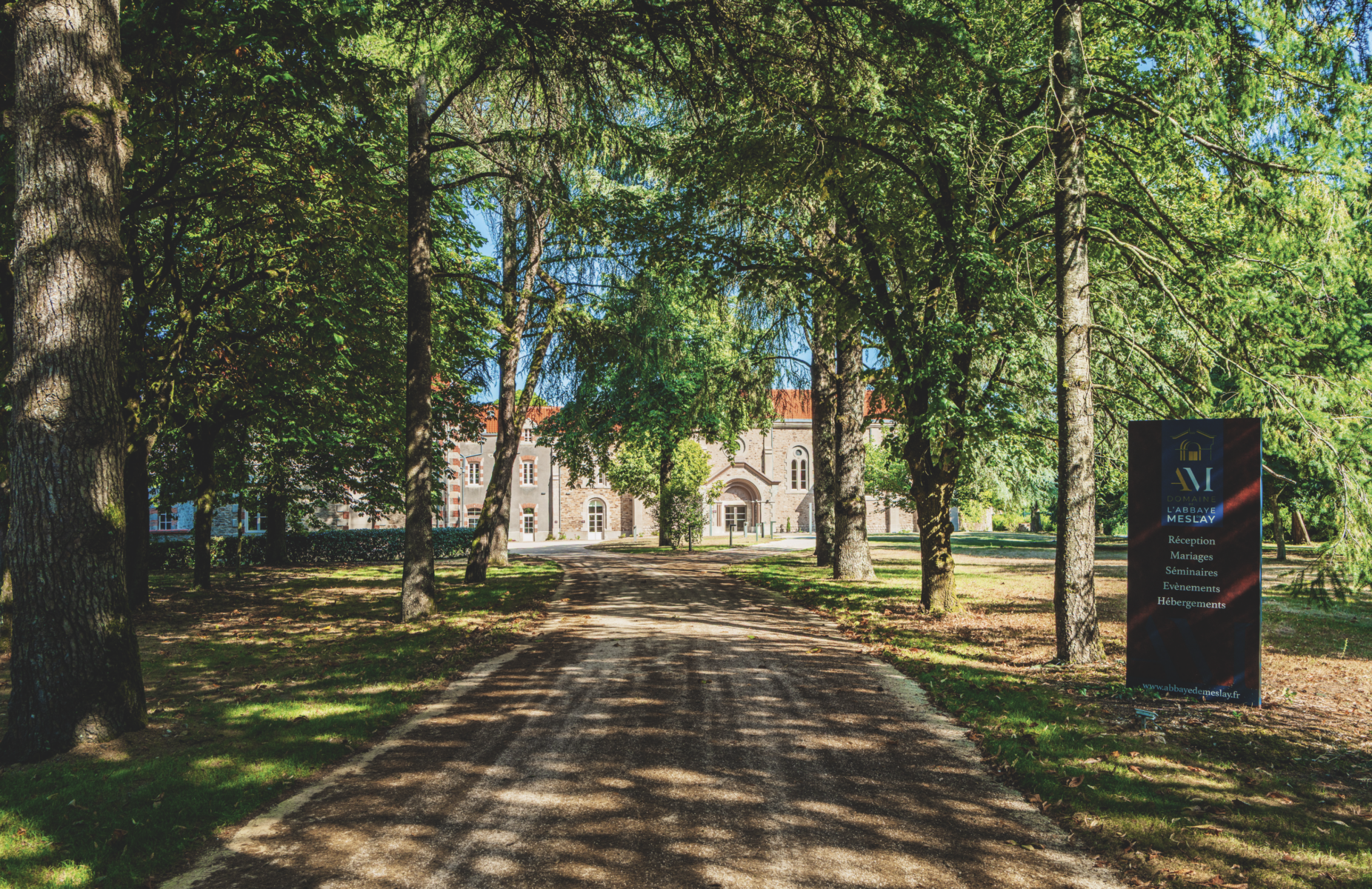 Un chemin bordé d'arbres mène au bâtiment historique en briques de la Grimpe Atlantique Ouest, aux fenêtres cintrées et au toit rouge. Un panneau sur la droite indique les coordonnées de la réception, du restaurant et de l'hébergement, tandis que la lumière du soleil filtre à travers la canopée verte. - Édition Offset 5 (Partenaire de votre communication locale en Vendée)