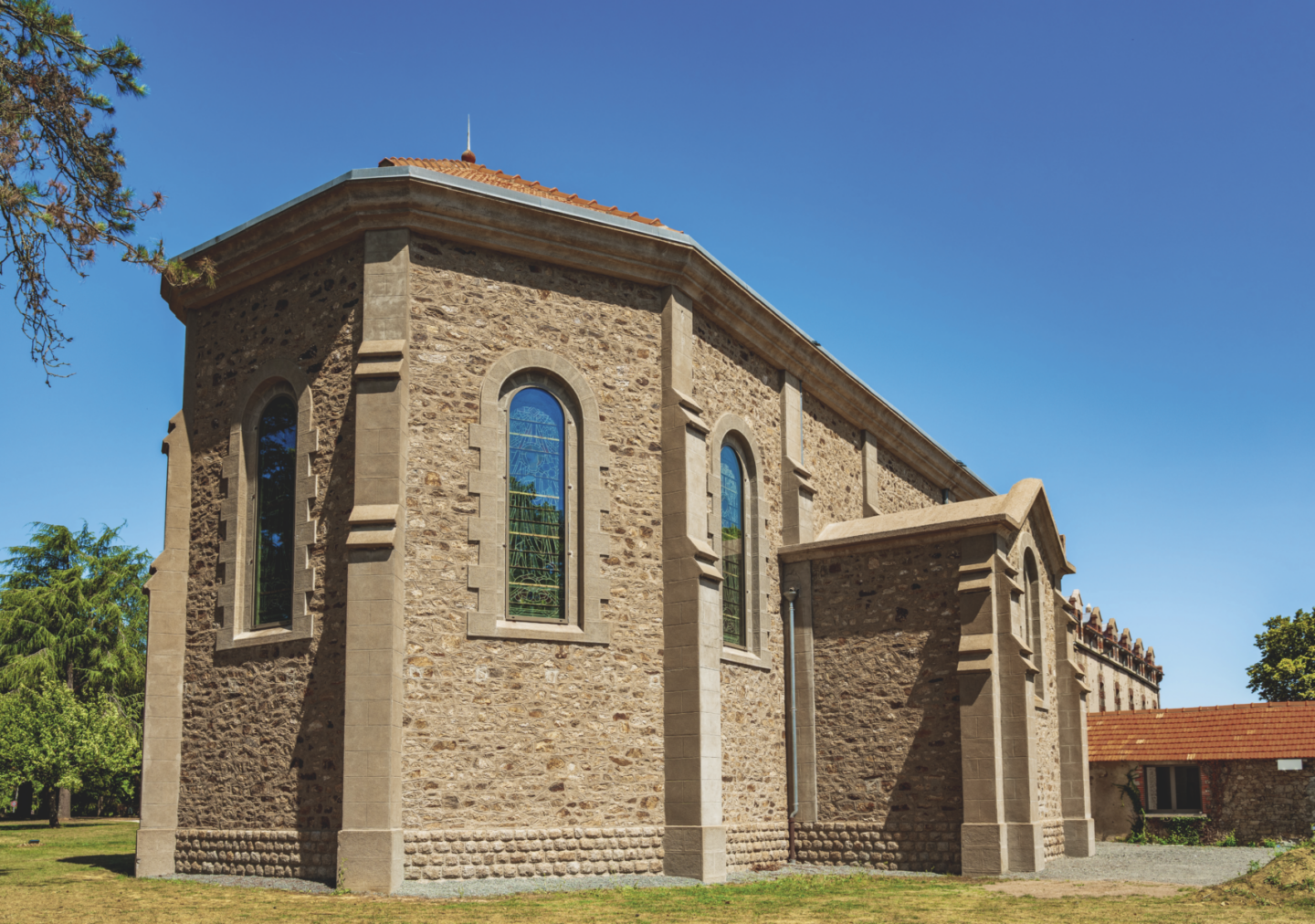 Église en pierre avec vitraux cintrés, toit de tuiles rouges et herbe environnante sous un ciel bleu clair. Des arbres et un autre bâtiment à toit rouge de la Grimpe Atlantique Ouest sont visibles à l'arrière-plan. - Édition Offset 5 (Partenaire de votre communication locale en Vendée)