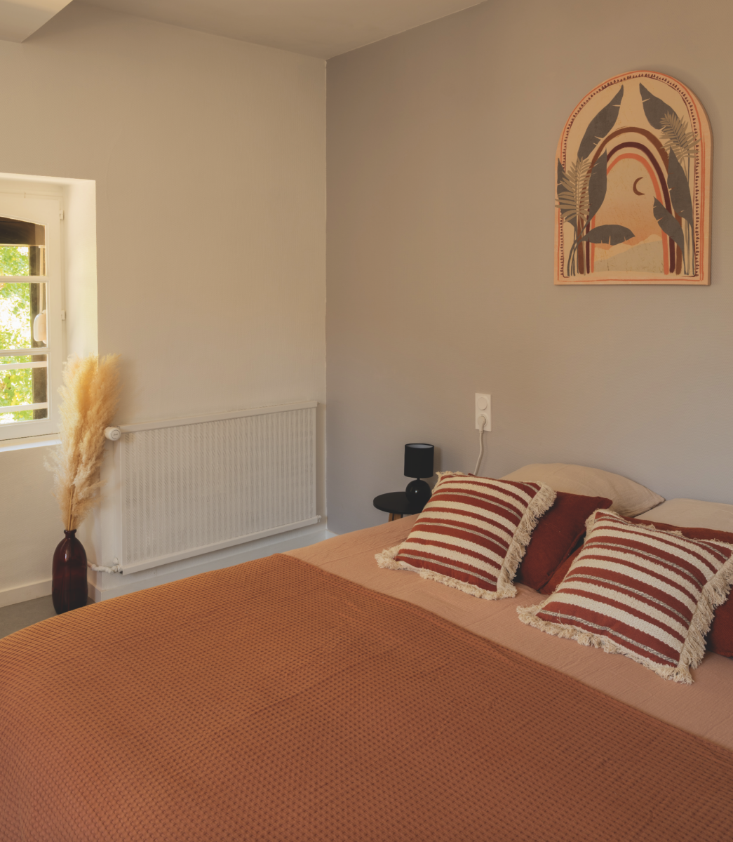 Une chambre douillette avec un couvre-lit orange, deux oreillers rayés, une petite table de nuit noire et une tenture murale Grimpe Atlantique Ouest avec des formes abstraites et des feuilles au-dessus du lit, ainsi qu'un vase avec de l'herbe de la pampa près de la fenêtre. - Édition Offset 5 (Partenaire de votre communication locale en Vendée)