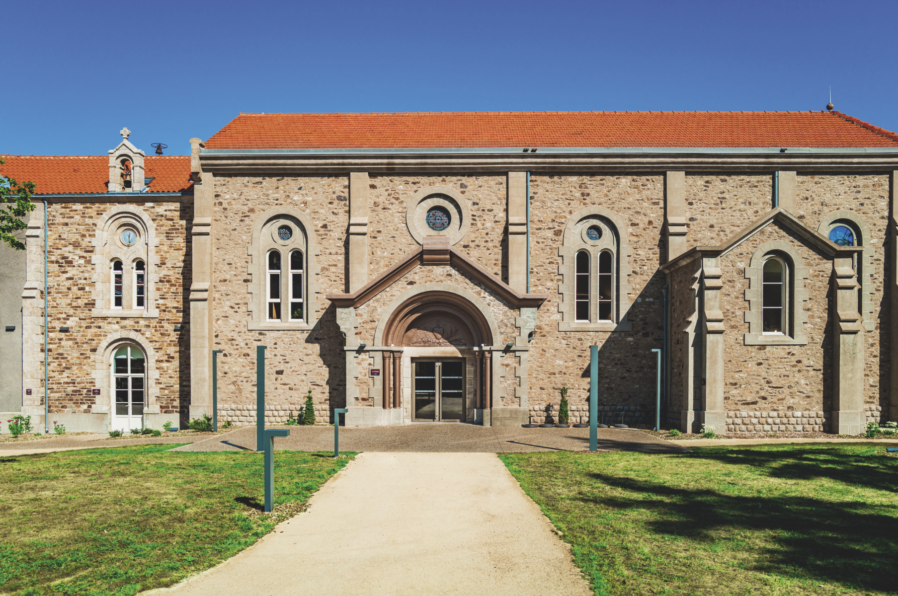 Un bâtiment historique en pierre avec des fenêtres cintrées, un toit en tuiles rouges et une entrée centrale cintrée, se trouve au milieu de pelouses et d'allées sous un ciel bleu clair - le cadre idéal pour les réunions du Grimpe Atlantique Ouest. - Édition Offset 5 (Partenaire de votre communication locale en Vendée)