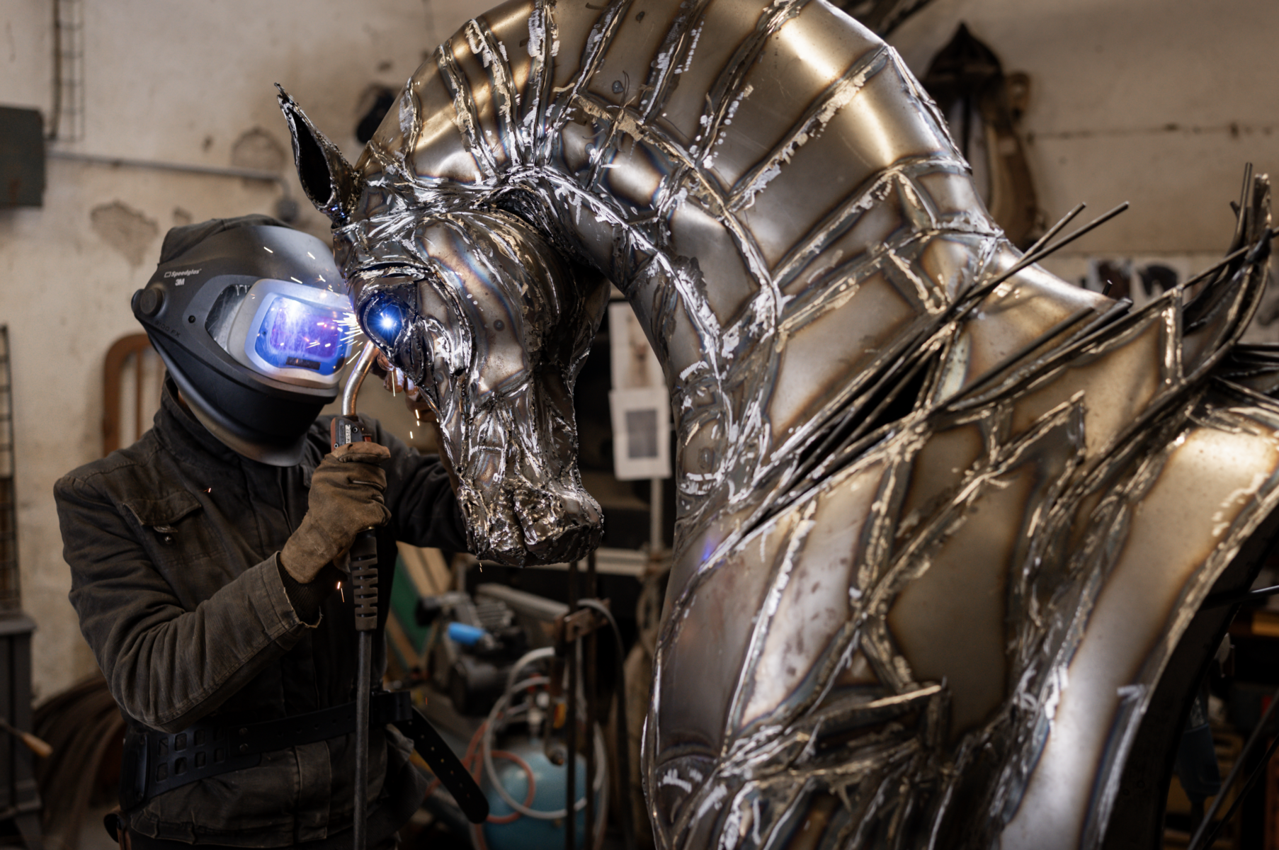 Dans un atelier, une personne portant un équipement de protection soude des détails sur une grande sculpture métallique de cheval aux yeux bleus brillants, afin de la préparer pour Les nocturnes du Château de Talmont. - Édition Offset 5 (Partenaire de votre communication locale en Vendée)