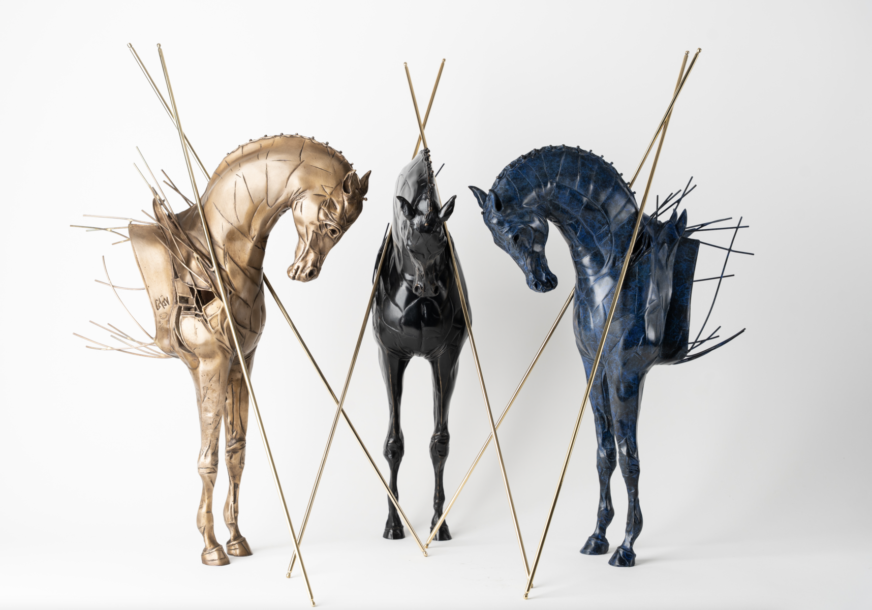 Trois sculptures de chevaux stylisés - or, noir et bleu - se côtoient, rehaussées de tiges et de pointes métalliques, évoquant l'ambiance mystique des nocturnes du Château de Talmont. Plusieurs tiges traversent le fond blanc au premier plan. - Édition Offset 5 (Partenaire de votre communication locale en Vendée)