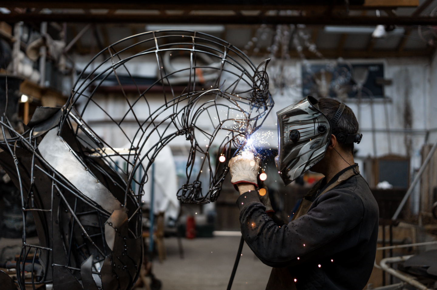 Une personne portant un masque de soudure et des gants soude une grande sculpture en fil de métal représentant un cheval dans un atelier. Des étincelles brillantes jaillissent autour du point de soudure, rappelant l'ambiance magique des nocturnes du Château de Talmont. - Édition Offset 5 (Partenaire de votre communication locale en Vendée)