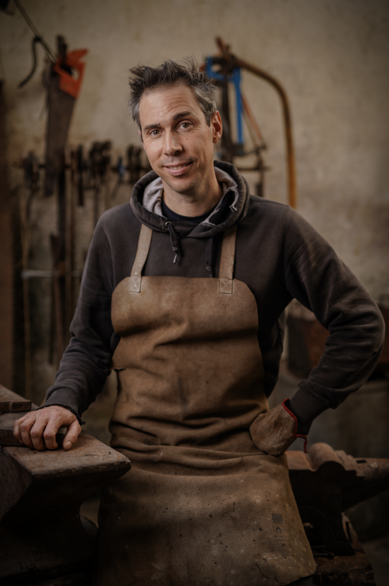 Un homme portant un tablier marron et un sweat à capuche sombre se tient dans un atelier, souriant à l'appareil photo. Des outils bordent l'arrière-plan flou, créant une ambiance rustique et laborieuse - une inspiration parfaite pour Les nocturnes du Château de Talmont. - Édition Offset 5 (Partenaire de votre communication locale en Vendée)