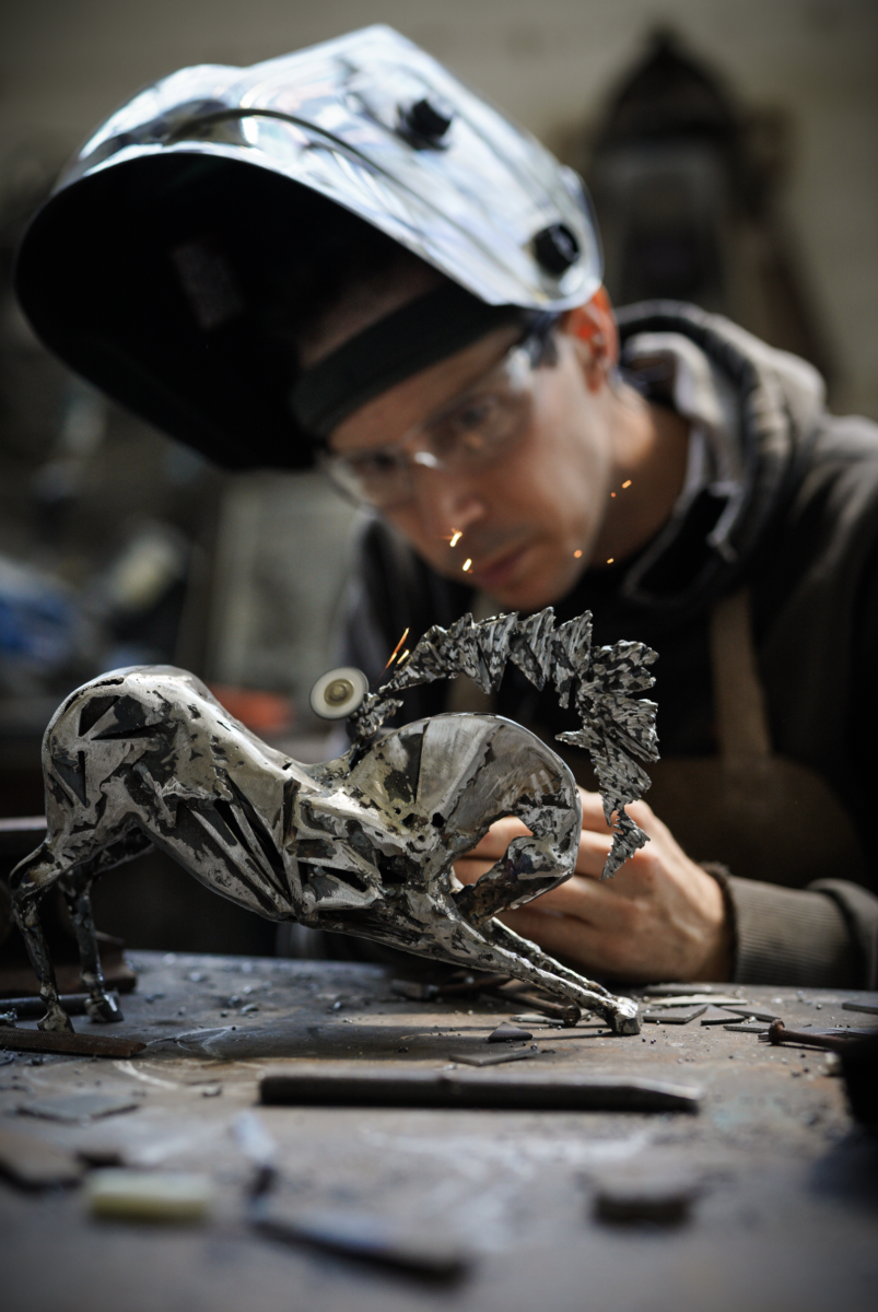 Une personne portant des lunettes de sécurité et un masque de soudure travaille sur une sculpture métallique représentant un cheval, à l'aide d'un outil qui produit des étincelles. Des outils et des fragments de métal sont éparpillés sur l'établi, rappelant la créativité des nocturnes du Château de Talmont. - Édition Offset 5 (Partenaire de votre communication locale en Vendée)