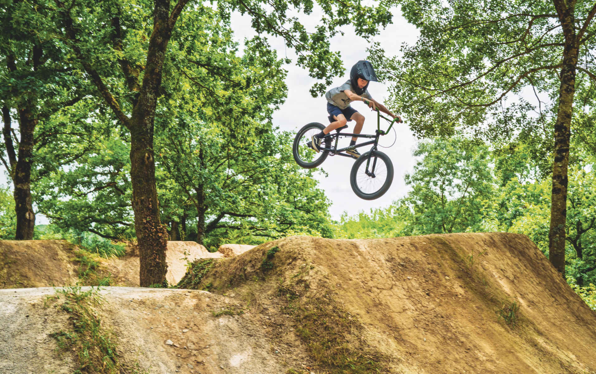 Une personne portant un casque saute à vélo BMX sur une rampe en terre dans un parc boisé près du Manoir de la Mortière, entouré d'arbres verts et feuillus. - Édition Offset 5 (Partenaire de votre communication locale en Vendée)