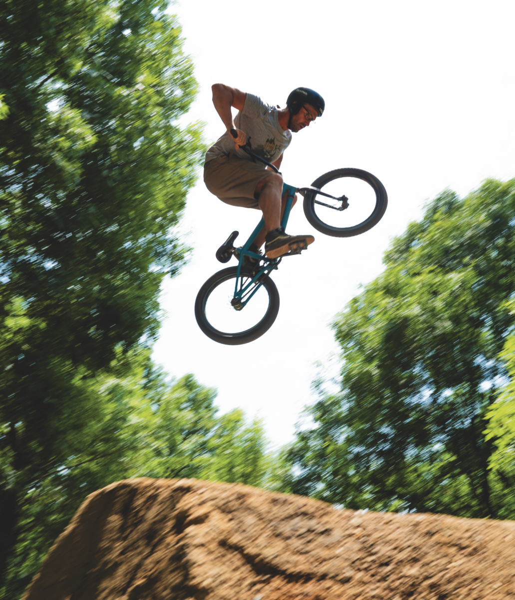 Une personne portant un casque effectue un saut sur un vélo BMX au-dessus d'une rampe en terre battue au Manoir de la Mortière, entouré d'arbres verts sous un ciel lumineux. - Édition Offset 5 (Partenaire de votre communication locale en Vendée)