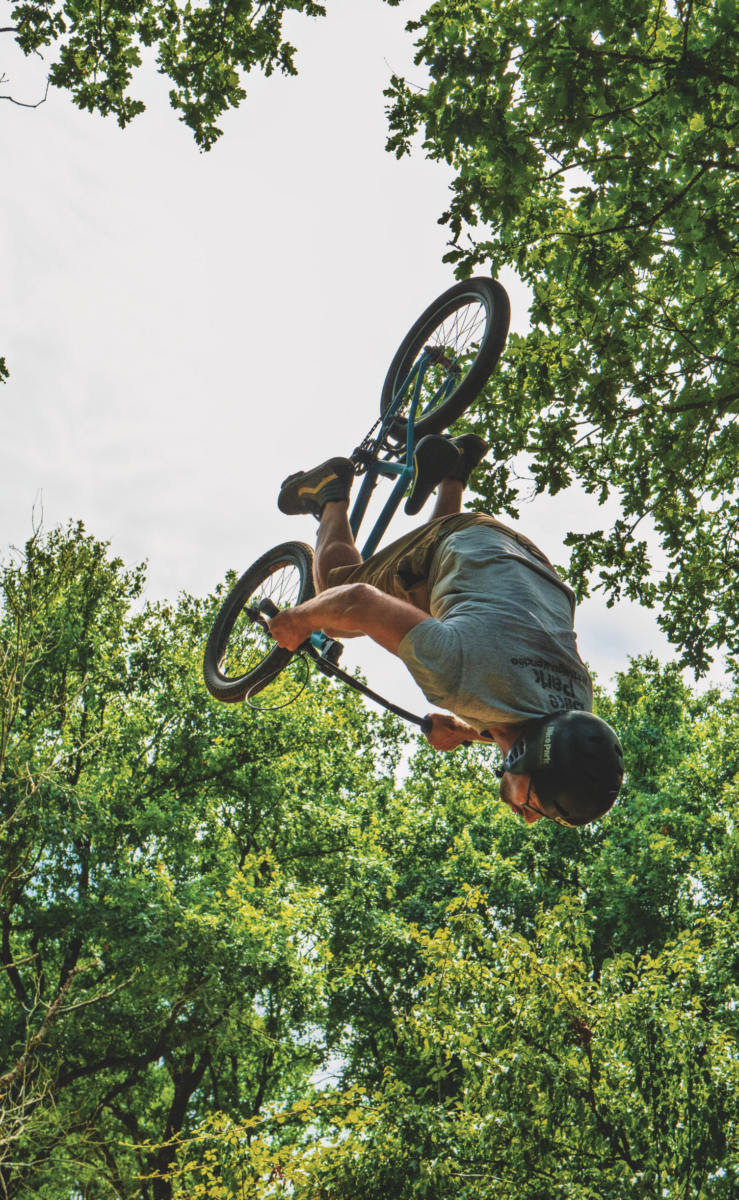 Un pilote de BMX portant un casque effectue une figure aérienne à l'envers au-dessus d'arbres verts, avec le ciel lumineux et Le Manoir de la Mortière en arrière-plan. - Édition Offset 5 (Partenaire de votre communication locale en Vendée)