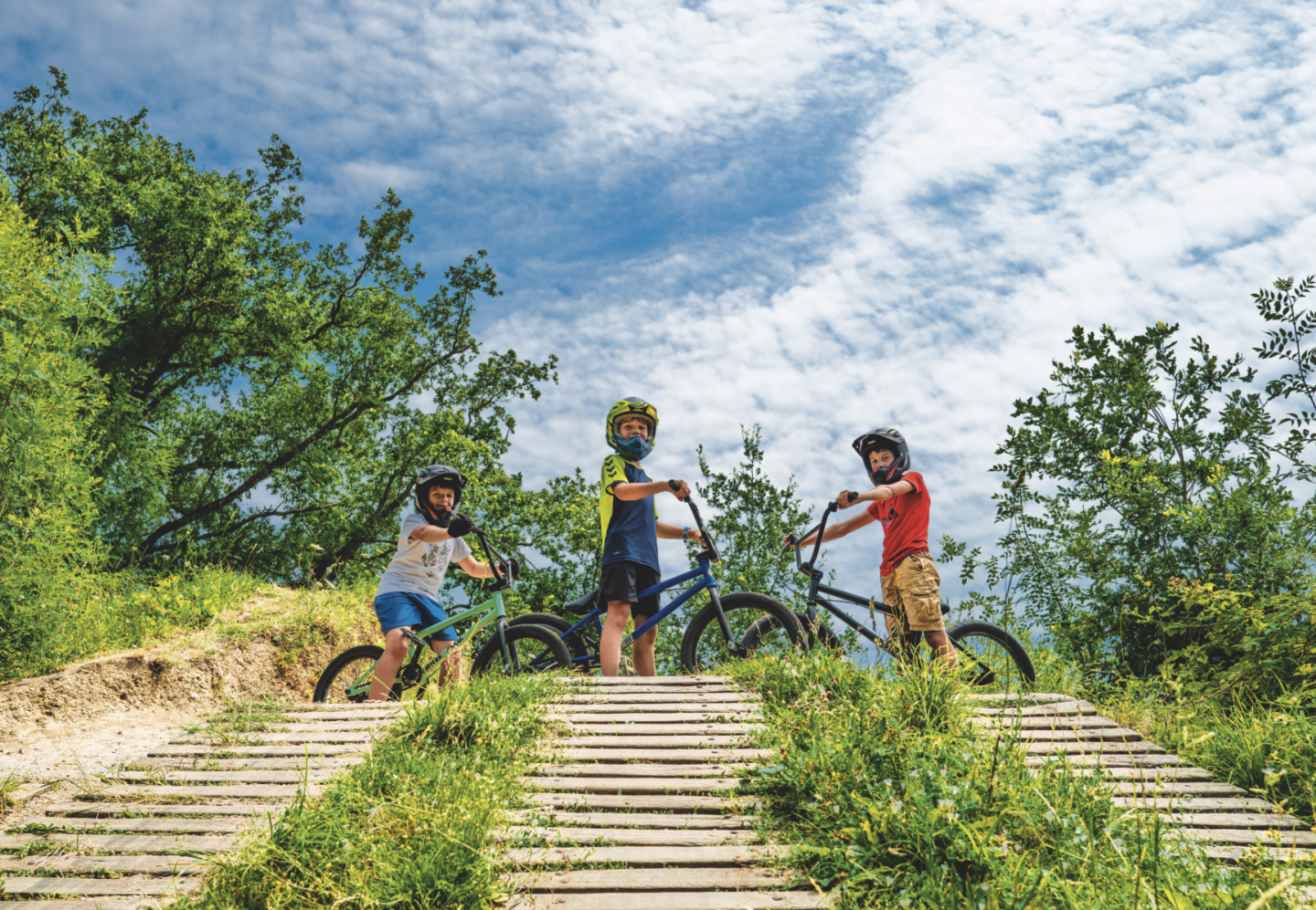 Trois enfants portant des casques sont debout avec leurs vélos sur une rampe en bois au Manoir de la Mortière, entourés d'arbres verts sous un ciel bleu partiellement nuageux. Ils semblent prêts à dévaler la pente ou à faire la course. - Édition Offset 5 (Partenaire de votre communication locale en Vendée)