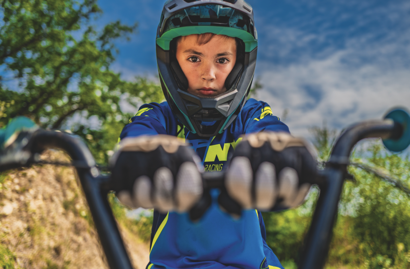Un jeune garçon portant un casque et une tenue de cycliste bleue tient le guidon de son vélo, regardant devant lui avec détermination, avec des arbres et un ciel partiellement nuageux en arrière-plan, près du Manoir de la Mortière. - Édition Offset 5 (Partenaire de votre communication locale en Vendée)