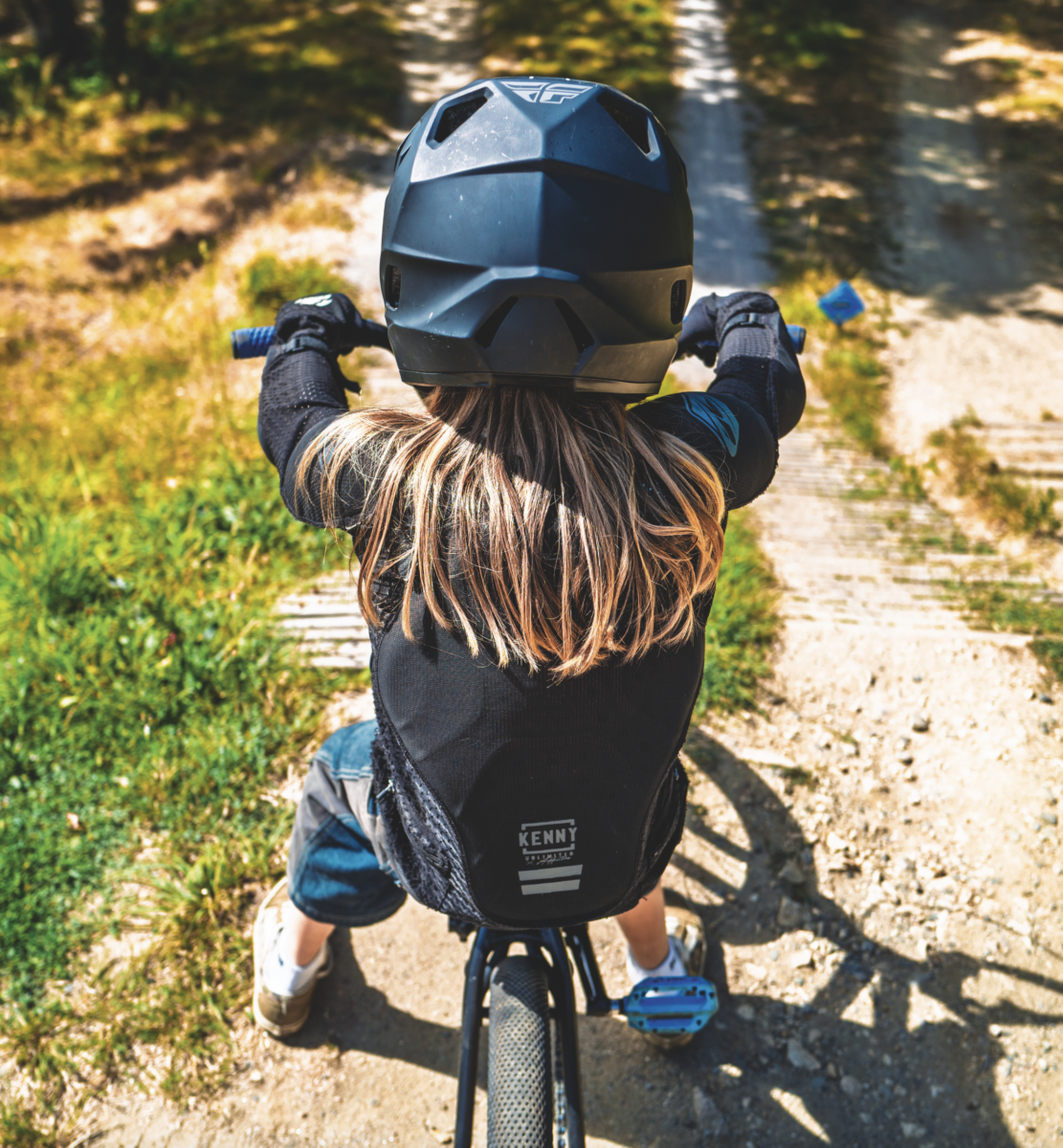 Un enfant portant un casque noir et un équipement de protection roule à vélo sur un chemin de terre au Manoir de la Mortière, entouré d'herbe et d'arbres, vu de l'arrière. - Édition Offset 5 (Partenaire de votre communication locale en Vendée)