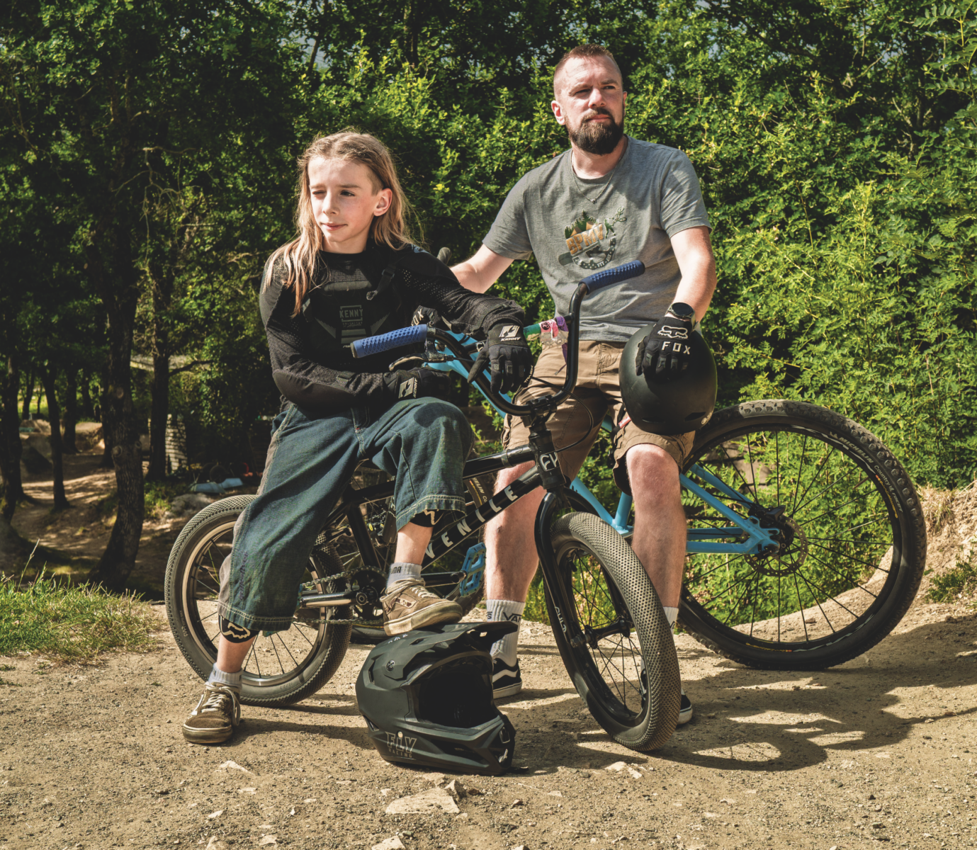 Un jeune et un adulte à vélo font une pause sur un chemin de terre dans une zone boisée et ensoleillée près du Manoir de la Mortière. Tous deux portent des vêtements décontractés ; les casques reposent sur le sol tandis qu'ils regardent au loin d'un air pensif. - Édition Offset 5 (Partenaire de votre communication locale en Vendée)