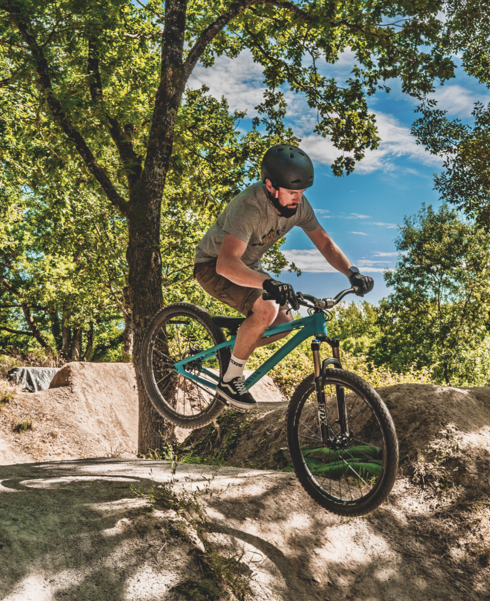 Un homme portant un casque et des gants roule en VTT sur un tremplin en terre battue dans la forêt ensoleillée près du Manoir de la Mortière, entouré d'arbres verts et sous un ciel bleu clair. - Édition Offset 5 (Partenaire de votre communication locale en Vendée)