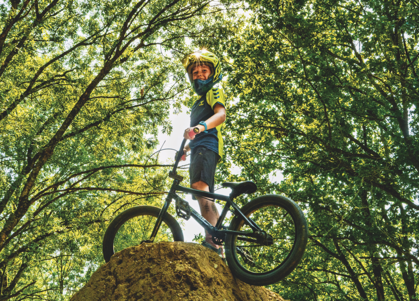Un jeune garçon portant un casque se tient sur un monticule de terre avec son vélo BMX au Manoir de la Mortière, entouré d'arbres et de la lumière du soleil filtrant à travers les feuilles. Il sourit avec assurance tout en regardant l'appareil photo. - Édition Offset 5 (Partenaire de votre communication locale en Vendée)