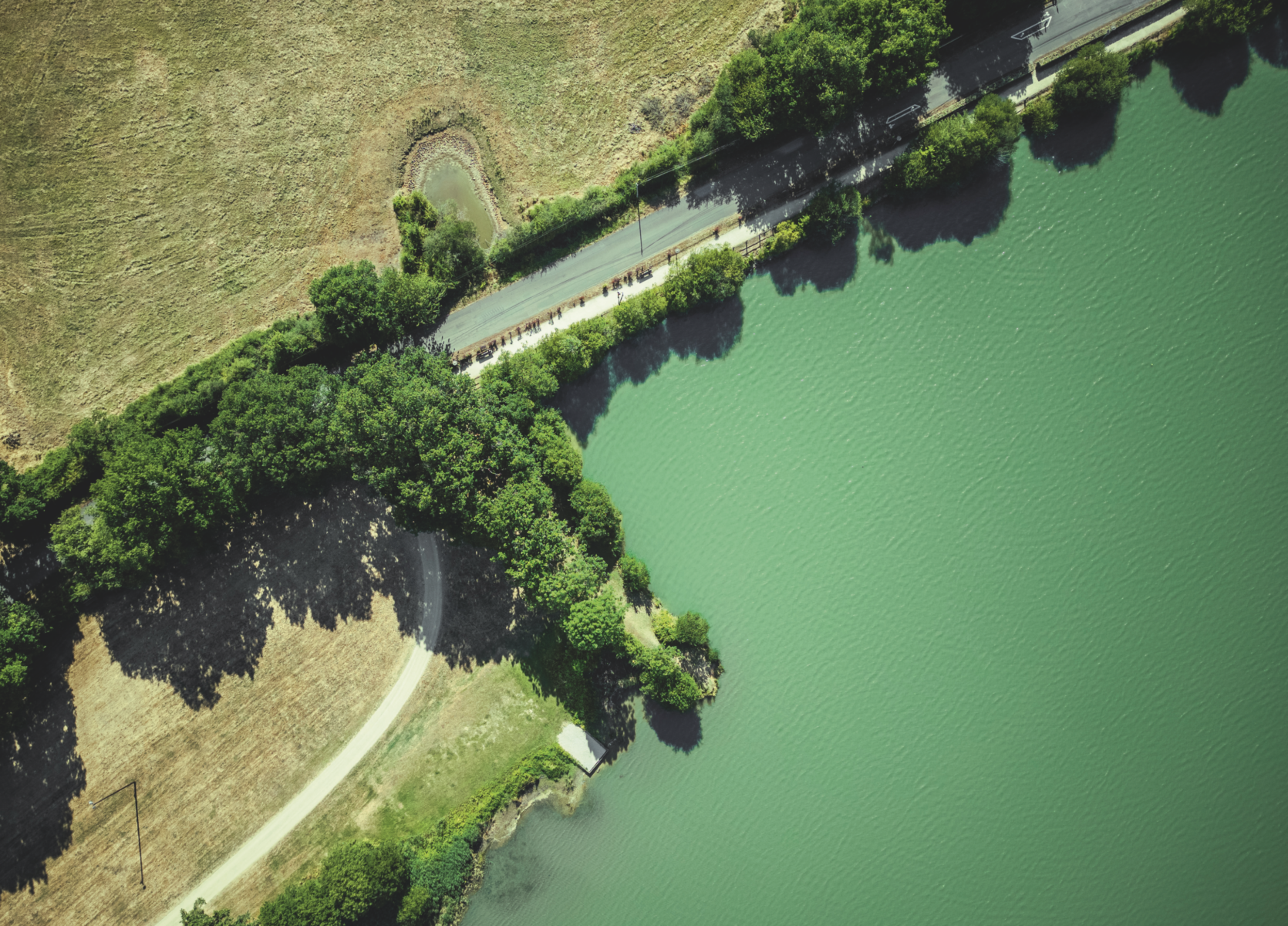 Vue aérienne de la route sinueuse du Veillon bordée d'arbres, passant entre une rivière bleu verdâtre et un champ sec et herbeux par une journée ensoleillée. Les ombres des arbres s'étendent sur la route et le champ. - Édition Offset 5 (Partenaire de votre communication locale en Vendée)