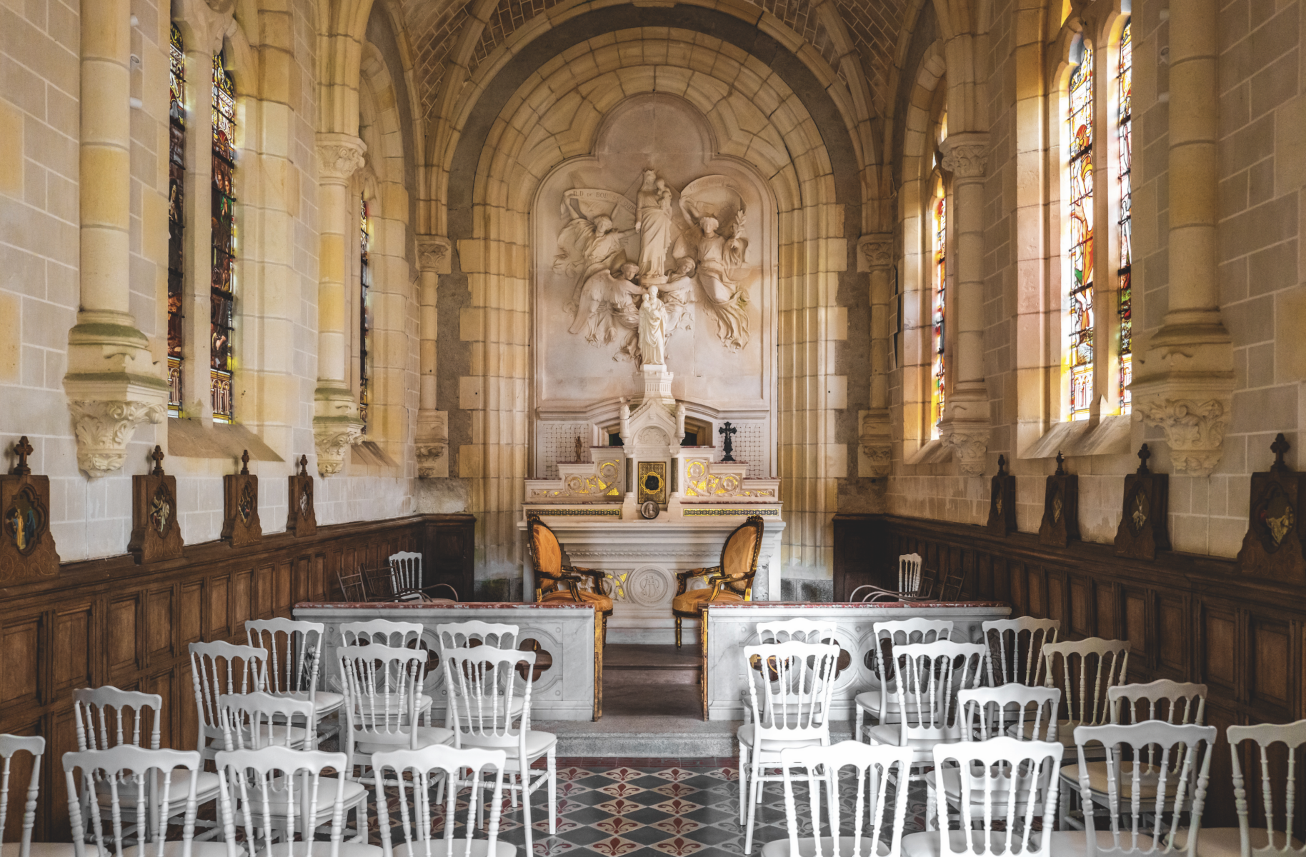 Intérieur d'une petite chapelle à Montaigu-Vendée avec des rangées de chaises blanches face à un autel orné d'une grande sculpture flanquée de vitraux et d'arcs décoratifs en pierre. Deux chaises rembourrées sont placées près de l'autel. - Édition Offset 5 (Partenaire de votre communication locale en Vendée)