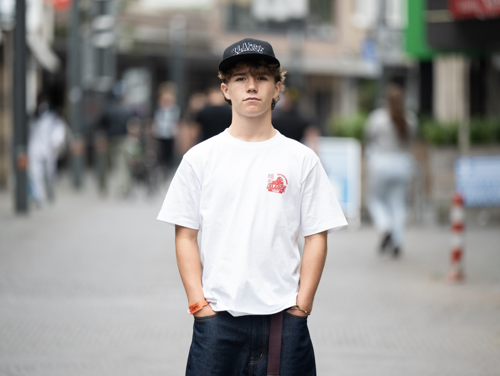 Un jeune se tient dans une rue de la ville, vêtu d'une casquette noire, d'un tee-shirt blanc avec un motif rouge et d'un pantalon sombre. Les mains dans les poches, la scène animée évoque des souvenirs de lieux comme l'Abbaye de Meslay. - Édition Offset 5 (Partenaire de votre communication locale en Vendée)