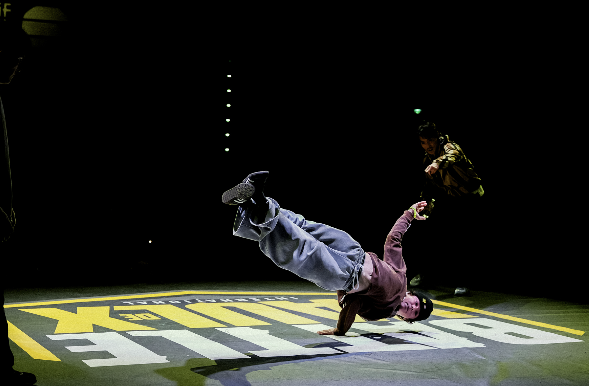 Un breakdancer vêtu d'un sweat-shirt bordeaux et d'un jean exécute un mouvement dynamique à une main sur la scène de l'Abbaye de Meslay lors d'un battle de breakdance, sous le regard d'un autre danseur en arrière-plan. Les mots BATTLE et BUJIK sont affichés sur le sol. - Édition Offset 5 (Partenaire de votre communication locale en Vendée)