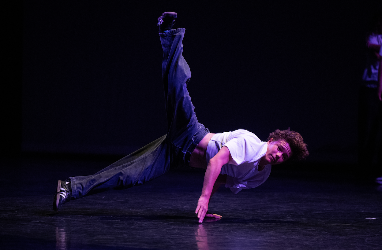 Ruben Boisseleau, vêtu d'un t-shirt blanc et d'un jean, se tient en équilibre sur une main tout en exécutant un mouvement dynamique de breakdance, les jambes tendues et la tête tournée vers l'appareil photo, sur une scène sombre. - Édition Offset 5 (Partenaire de votre communication locale en Vendée)