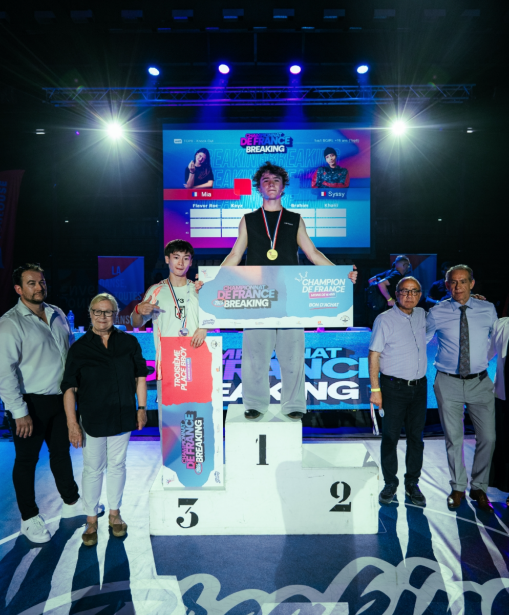 Un jeune athlète se tient sur un podium en première place, portant une médaille. Un autre athlète et Ruben Boisseleau se tiennent sur le podium de la deuxième place. Quatre adultes sont à leurs côtés, avec une grande toile de fond et des lumières colorées derrière eux. - Édition Offset 5 (Partenaire de votre communication locale en Vendée)